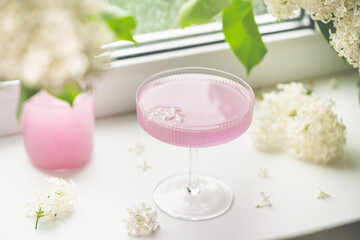 Obraz na płótnie Canvas Fresh pink non alcoholic cocktail in glass with lilac flowers against window background.