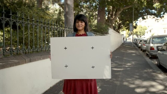 Female activist holding a white banner while standing next to a park outdoors. Confident woman smiling at the camera while holding a blank placard.
