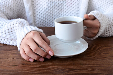 Close-up of girl's hands in a warm knitted white sweater holding cup of tea on  wooden table. Winter holidays and lifestyle concept