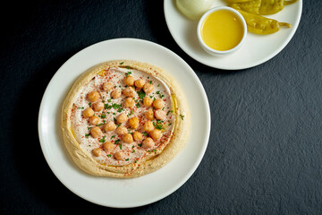 Hummus with olive oil, tahini and salted vegetables in a white plate on a black background