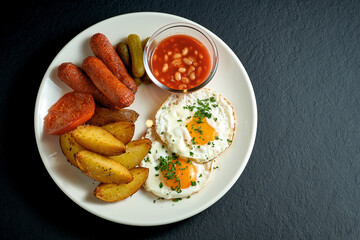 English breakfast - scrambled eggs with potatoes, beans, sausages in a white plate on a black background