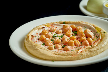 Hummus with olive oil, tahini and salted vegetables in a white plate on a black background
