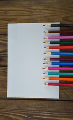 Colored pencils lie on a wooden table along a white sheet of paper. Blank white sheet of paper and pencils on a wooden table. Wooden colored pencils and a sheet of paper, top view