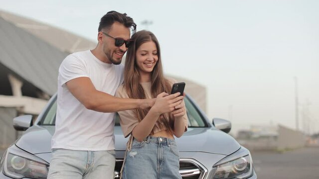 A man and a girl are standing near a car. They are smiling and watching something on their smartphone. They're talking. 4K