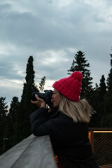 A blonde girl with hat taking a landscape photo in park. Side profile photo. There are pine trees behind her. Winter nature concept. Hobby and leisure time activity.