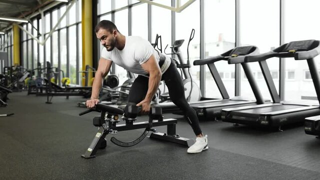 Male athlete doing dumbbell row exercise in gym indoors. Young muscular man, trains in sports club with sport bench. In the background is a large window and a row of treadmills. Sport concept.
