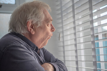 Senior man with shocked facial expression looking out of window blinds  - obrazy, fototapety, plakaty
