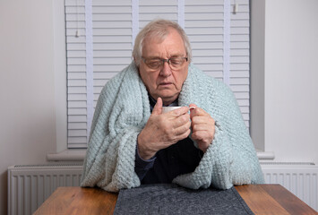 Senior man looking fed up holding a cup of coffee wrapped in a blanket 