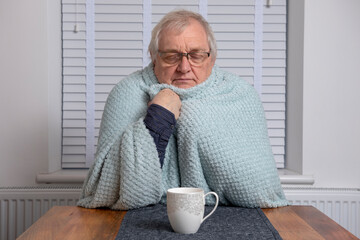 Cold senior man sitting with hot tea wrapped in a blanket 