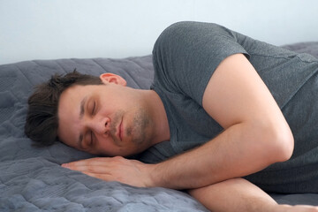 Portrait of young man is sleeping snoring in bed wearing in jeans and t-shirt after party. Man with morning hangover at home. He sleeps on unmade bed. Life of a person with bad habits, alcoholism.