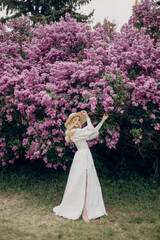 Obraz na płótnie Canvas Young blonde girl in a straw hat and in a white wedding dress with an open top near the lilac bushes
