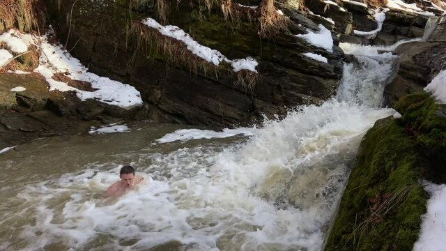 a caucasian man with a naked torso walks into an ice-hole on a river in a waterfall into icy water in winter. raises his hands to heaven. Epiphany in winter in the river. overall plan