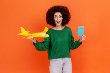 Surprised woman with Afro hairstyle in green casual sweater standing with plane and passport in hands, looking at camera, shocked with low prices. Indoor studio shot isolated on orange background.