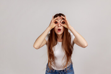 Portrait of amazed curious woman spying looking at camera through fingers imitating binoculars,...