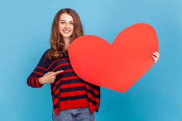 Young adult woman wearing striped casual style sweater, standing pointing at big red heart, looking at camera with smile and happy expression. Indoor studio shot isolated on blue background.