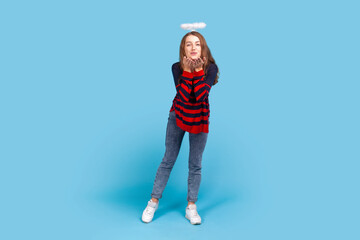 Full length of romantic angelic woman wearing striped sweater and nimb over head, standing and sending air kisses, looking at camera, falling in love. Indoor studio shot isolated on blue background.