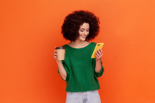 Good looking woman with Afro hairstyle wearing green casual style sweater drinking coffee and using smart phone , browsing internet while having break. Indoor studio shot isolated on orange background