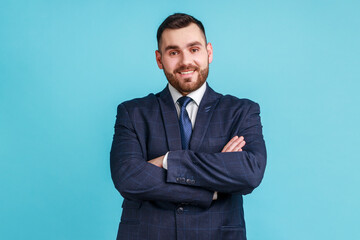 Young happy successful businessman wearing official style suit posing with happy confident expression, looking at camera, keeping arms folded. Indoor studio shot isolated on blue background.