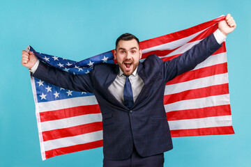 Extremely happy man with beard wearing official style suit holding USA flag and screaming happily, looking at camera, celebrating national holiday. Indoor studio shot isolated on blue background.