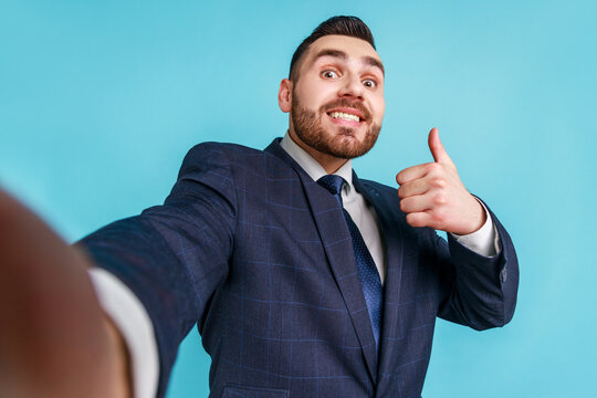 Smiling man with pleasant appearance wearing official style suit taking selfie, looking at camera and showing thumb up POV, point of view of photo. Indoor studio shot isolated on blue background.