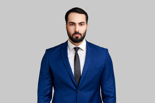 Self confident bearded male looking at camera with serious expression, unsmiling determined business man, wearing official style suit. Indoor studio shot isolated on gray background.