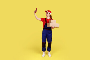 Full length portrait of courier woman standing with two packages in hands, taking selfie for delivery service social network, wearing overalls and cap. Indoor studio shot isolated on yellow background
