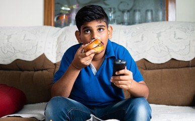 indian teenger kid watching tv or television by eating unhealthy food at home - concept of lesieur...