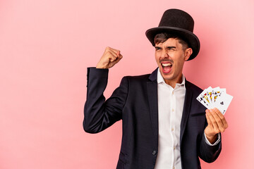 Young caucasian wizard man holding a magic cards isolated on white background raising fist after a...