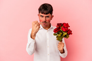 Young mixed race man holding a bouquet of flowers isolated on pink background showing fist to camera, aggressive facial expression.