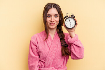 Young caucasian woman wearing a bathrobe holding a alarm clock isolated on yellow background happy, smiling and cheerful.