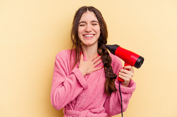 Young woman holding a hairdryer isolated on yellow background laughs out loudly keeping hand on chest.