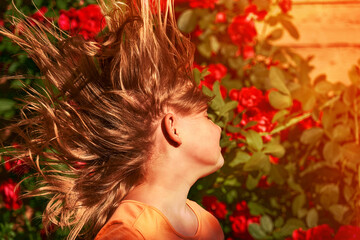 Naklejka premium A child's long hair grows in strong winds. Girl's hairstyle close-up on a background of roses in the garden. A child plays and indulges outdoor on a summer day.