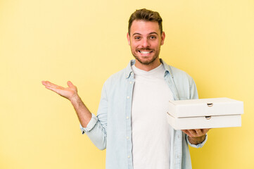 Young caucasian man holding a pizzas isolated on yellow background showing a copy space on a palm and holding another hand on waist.