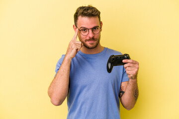 Young caucasian man holding a game controller isolated on yellow background pointing temple with finger, thinking, focused on a task.