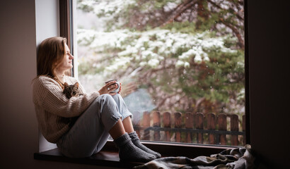 Young cheerful girl sitting at home on the windowsill in a warm sweater drinking coffee with gray kitten in her arms
