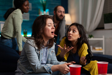 Obraz premium Nevours upset women sitting on sofa playing video games using gaming joystick losing online competition. Group of multi-ethnic friends hanging out together having fun during wekeend party at home