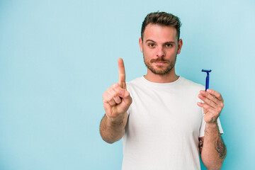 Young caucasian man shaving his beard isolated on blue background showing number one with finger.