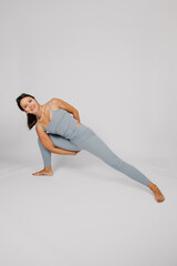 Sporty young woman does yoga. Concept of healthy lifestyle, brunette in gray fitness suit stands in yoga pose on isolated gray background, smiles and shows her flexibility. 
