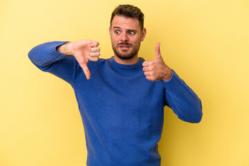 Young caucasian man isolated on yellow background showing thumbs up and thumbs down, difficult...