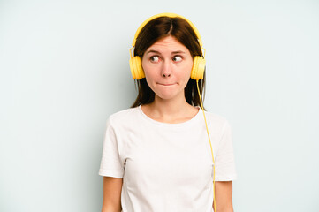 Young English woman listening to music isolated on blue background confused, feels doubtful and unsure.