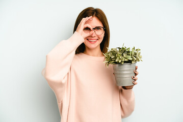 Young English woman holding a plant isolated on blue background excited keeping ok gesture on eye.