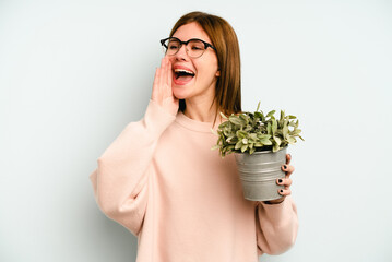 Young English woman holding a plant isolated on blue background shouting and holding palm near opened mouth.