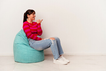 Young Argentinian woman sitting on a puff isolated on white background points with thumb finger away, laughing and carefree.