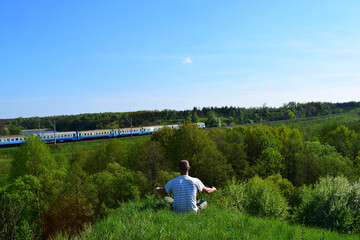A tourist sits on a hill in the green grass and enjoys the view. a man looks at a passing train. back view. nice spring weather. outdoors in summer. young man in nature