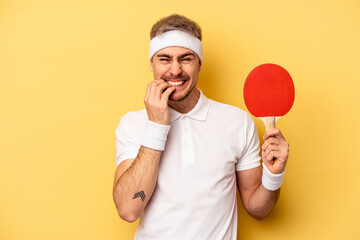 Young caucasian man holding ping pong rackets isolated on yellow background biting fingernails,...
