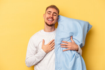 Young caucasian man wearing a pajama and holding pillow isolated on yellow background laughs out...