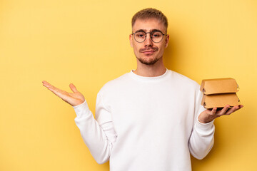 Young caucasian man holding a burger isolated on yellow background showing a copy space on a palm and holding another hand on waist.