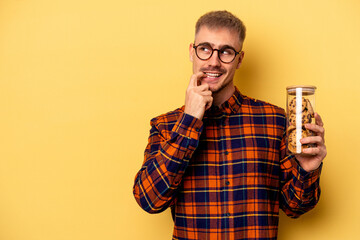Young caucasian man holding cookies jar isolated on yellow background relaxed thinking about something looking at a copy space.