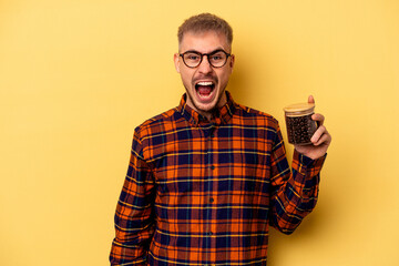 Young caucasian man holding coffee jar isolated on yellow background screaming very angry and...
