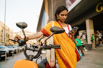 Young indian woman using cellphone while standing with scooter outdoors